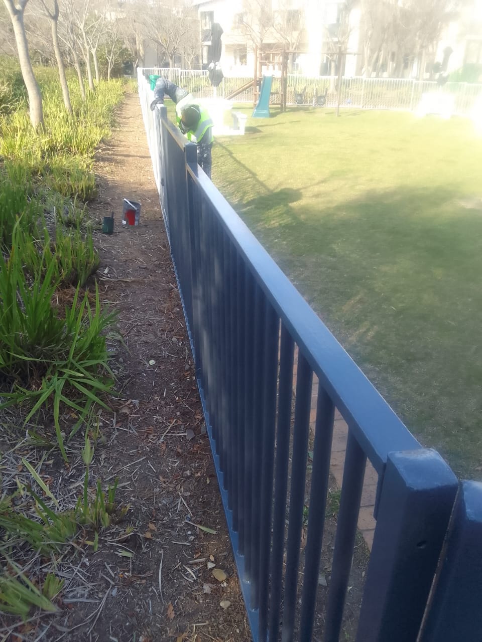 Long steel estate palisade fence being re-enamelled navy, playground behind